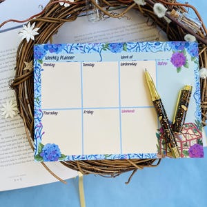 May include: A weekly planner with a floral design in shades of blue and purple. The planner has spaces for each day of the week, including Monday, Tuesday, Wednesday, Thursday, Friday, and Weekend. There is also a notes section. Two gold pens are lying on top of the planner.