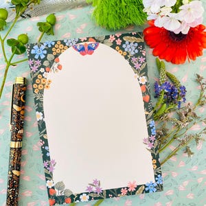 以下が含まれることがあります： 蝶、イチゴ、さまざまな花が描かれた花のボーダーが付いた、空白の文房具カード。 天体のデザインの装飾的なペンがカードの隣にあります。 背景は、明るい緑色の花柄です。