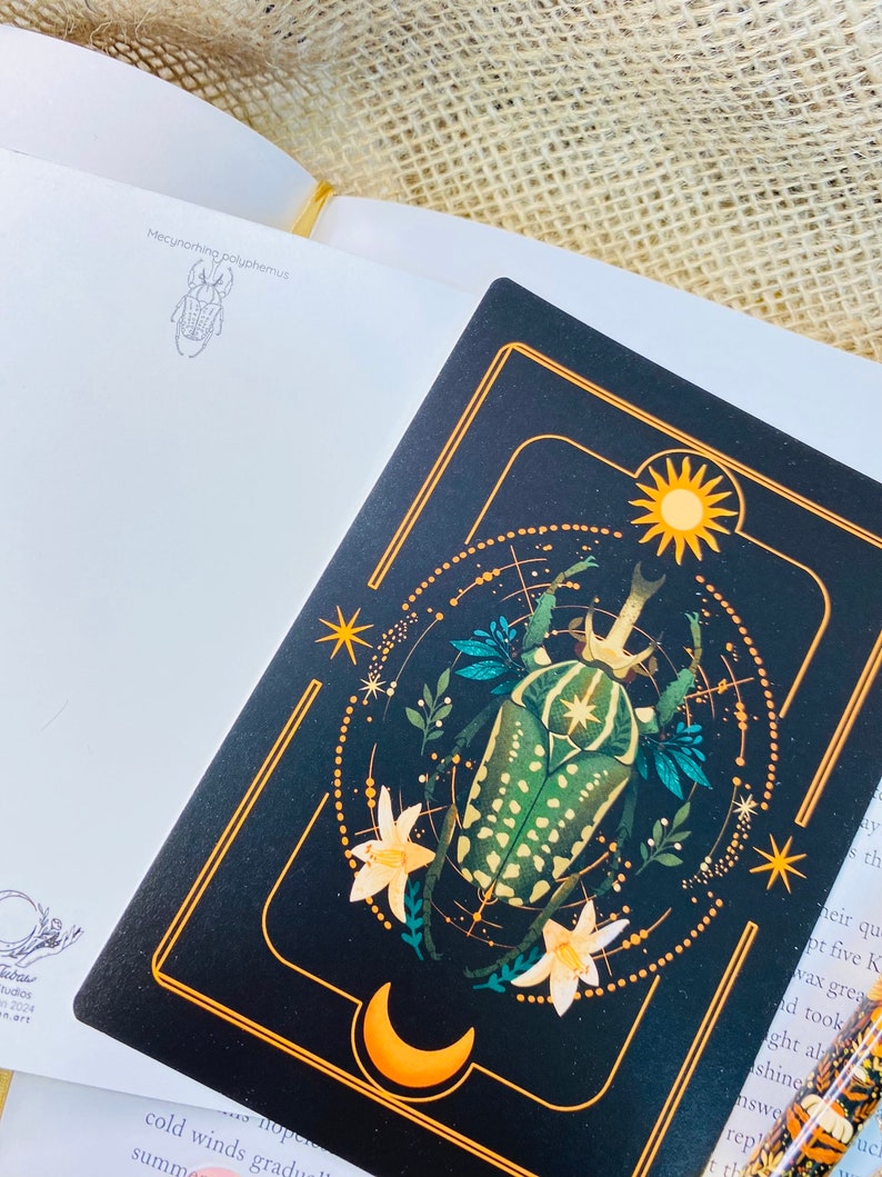 May include: A black and gold illustrated card with a green and yellow beetle, flowers, a crescent moon, and a sun. The text "Mecynorhina polyphemus" is at the top of the card.