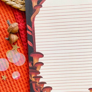May include: A lined notepad with a black border featuring a mushroom and branch design in shades of orange and brown. The notepad is on a textured orange surface with acorns and star-shaped confetti.