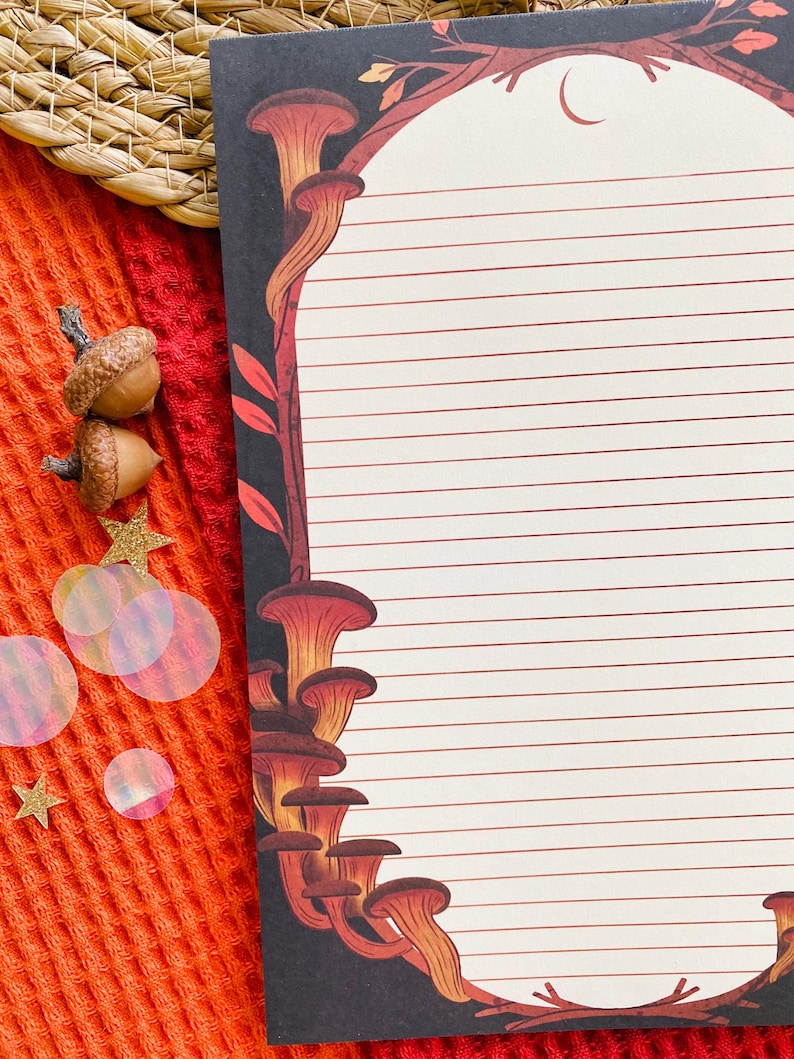 May include: A lined notepad with a black border featuring a mushroom and branch design in shades of orange and brown. The notepad is on a textured orange surface with acorns and star-shaped confetti.
