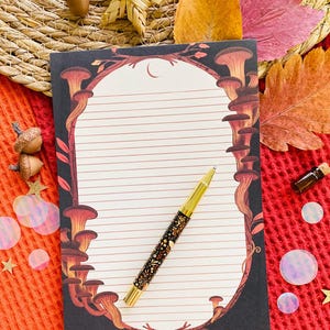 May include: A lined notepad with a mushroom illustration and a gold pen with a floral design. The notepad has a black border with brown and orange mushrooms. The pen is gold with black and gold floral details. The background includes fall leaves and decorative elements.