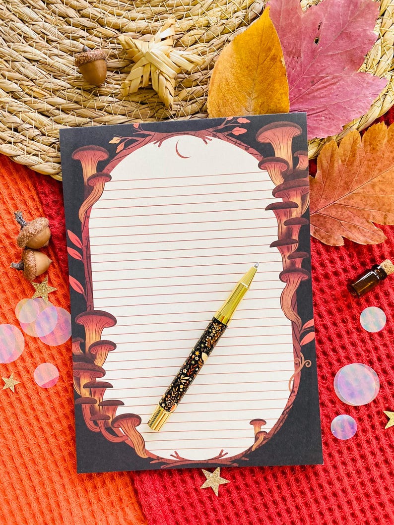 May include: A lined notepad with a mushroom illustration and a gold pen with a floral design. The notepad has a black border with brown and orange mushrooms. The pen is gold with black and gold floral details. The background includes fall leaves and decorative elements.
