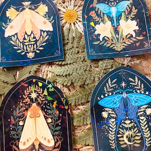 K&ouml;nnte beinhalten: Vier illustrierte Aufkleber mit Motten und Blumen auf dunkelblauem Hintergrund mit goldenen Akzenten. Jeder Aufkleber ist in Form eines Bogenfensters mit einem Halbmond am unteren Rand gestaltet.