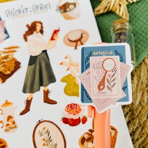 以下が含まれることがあります： 「botanical」という文字が書かれた青い本のステッカーシート。本の上に、花のイラストが描かれた虫眼鏡が置かれています。ステッカーシートには、茶色の髪をした女性が白いシャツと緑のスカートを着ていたり、キノコのイラストやキノコのイラストが描かれたティーカップなどがあります。
