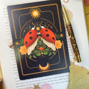 Peut inclure: Une carte noire avec une bordure dor&eacute;e pr&eacute;sente une illustration stylis&eacute;e d'une coccinelle avec des ailes, entour&eacute;e de fleurs et d'&eacute;toiles. Le soleil et la lune sont repr&eacute;sent&eacute;s dans les coins sup&eacute;rieurs. Le texte "THE BAMBOO-CUTTER AND THE MOON-CHILD" est imprim&eacute; en bas de la carte.