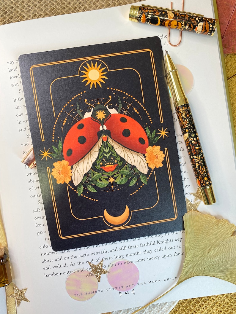 Peut inclure: Une carte noire avec une bordure dor&eacute;e pr&eacute;sente une illustration stylis&eacute;e d'une coccinelle avec des ailes, entour&eacute;e de fleurs et d'&eacute;toiles. Le soleil et la lune sont repr&eacute;sent&eacute;s dans les coins sup&eacute;rieurs. Le texte "THE BAMBOO-CUTTER AND THE MOON-CHILD" est imprim&eacute; en bas de la carte.