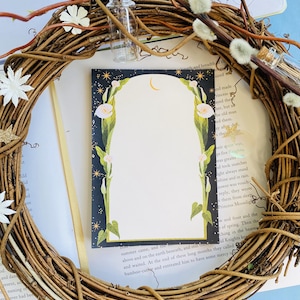 Könnte beinhalten: Eine leere Karte mit einem floralen Design mit weißen Blumen und einem zunehmenden Mond. Die Karte hat einen dunkelblauen Hintergrund mit weißen Sternen. Die Karte ist von einem Kranz aus Zweigen und weißen, flauschigen Knospen umgeben.