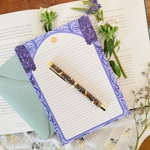 Könnte beinhalten: Ein Schreibwarenset mit einem linierten Notizblock mit lila Blumenrand, einem Stift mit braunem und goldenem Design und einem hellblauen Umschlag. Der Notizblock hat eine Sonnenillustration. Das Set wird auf einem offenen Buch mit Blumen präsentiert.