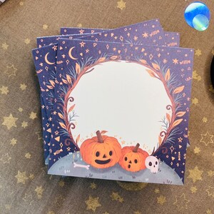 May include: A stack of square cards with a Halloween theme. Each card features a dark blue background with stars, moons, and a decorative border. The design includes orange pumpkins, a skull, and a bone. The cards are stacked on a gold-colored surface.