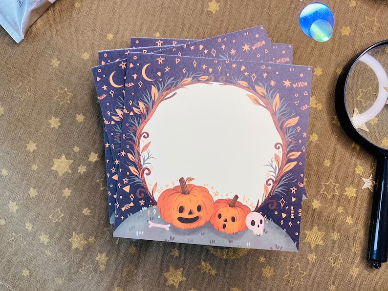 May include: A stack of square cards with a Halloween theme. Each card features a dark blue background with stars, moons, and a decorative border. The design includes orange pumpkins, a skull, and a bone. The cards are stacked on a gold-colored surface.