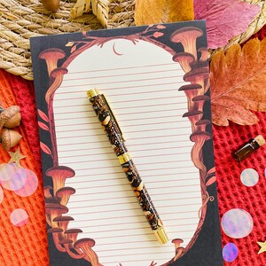 May include: A decorative pen and notepad set. The pen has a black barrel with a gold-colored clip and tip. The notepad features a mushroom design border and lined pages. The set is on a red textured surface with fall-themed decorations.