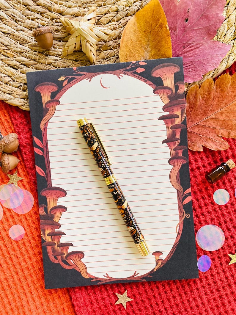 May include: A decorative pen and notepad set. The pen has a black barrel with a gold-colored clip and tip. The notepad features a mushroom design border and lined pages. The set is on a red textured surface with fall-themed decorations.