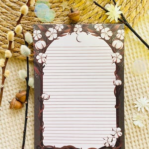 Pu&ograve; includere: Blocco note a righe con bordo floreale con fiori bianchi e tralci marroni. Il blocco note ha un bordo marrone scuro e un interno bianco con linee orizzontali. Elementi decorativi: ghiande e rametti.