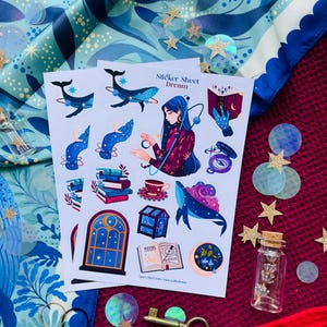 May include: Two sticker sheets featuring illustrations of whales, books, and a woman with long blue hair. The text "Sticker Sheet Dream" is visible. The stickers are on a red and blue patterned surface with decorative stars and confetti.