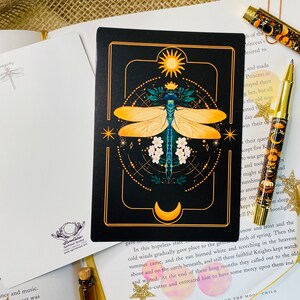 May include: A black card with a gold border features a detailed illustration of a dragonfly with yellow wings and a blue body. The dragonfly is surrounded by a geometric pattern with a sun and moon at the top and bottom. The card has the text "In this hopeless state..."