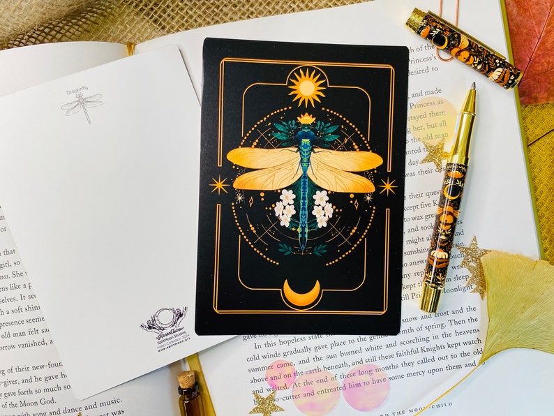 May include: A black card with a gold border features a detailed illustration of a dragonfly with yellow wings and a blue body. The dragonfly is surrounded by a geometric pattern with a sun and moon at the top and bottom. The card has the text "In this hopeless state..."