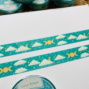 May include: Two rolls of teal washi tape with a moon and cloud design. The tape features a crescent moon, a triple moon symbol, and white clouds on a teal background with gold glitter. The text on the tape reads "Space Bird Crafts - Washi Tape - Fine"