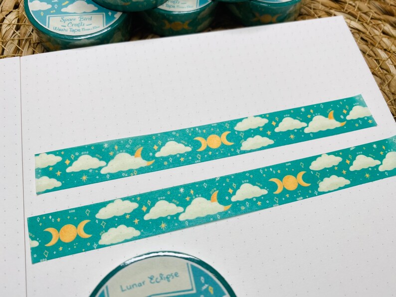 May include: Two rolls of teal washi tape with a moon and cloud design. The tape features a crescent moon, a triple moon symbol, and white clouds on a teal background with gold glitter. The text on the tape reads "Space Bird Crafts - Washi Tape - Fine"