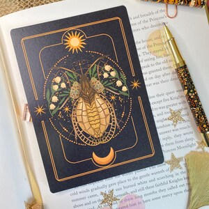 Peut inclure: Une carte de tarot noire et dor&eacute;e avec une illustration stylis&eacute;e d'un scarab&eacute;e &agrave; carapace dor&eacute;e et une lune croissante en arri&egrave;re-plan. La carte est bord&eacute;e d'&eacute;toiles blanches et d'un motif floral avec des fleurs blanches. Le texte "The Bamboo-Cutter and the Moon-Child" est imprim&eacute; en bas de la carte.