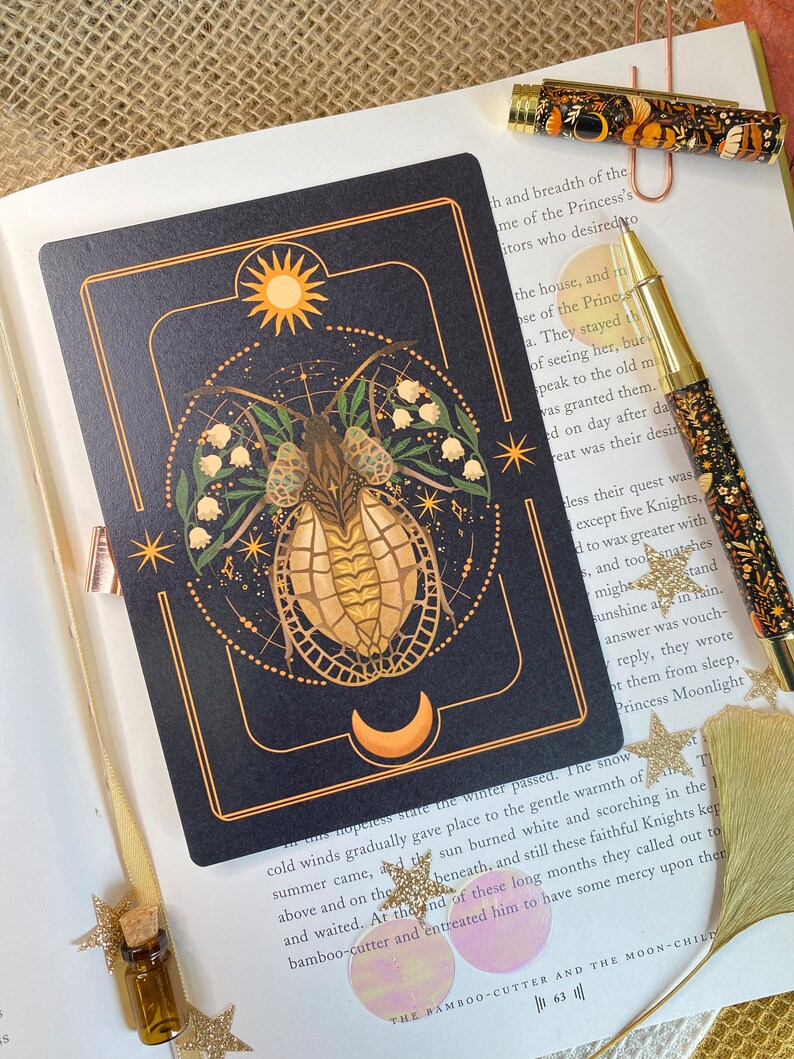Peut inclure: Une carte de tarot noire et dor&eacute;e avec une illustration stylis&eacute;e d'un scarab&eacute;e &agrave; carapace dor&eacute;e et une lune croissante en arri&egrave;re-plan. La carte est bord&eacute;e d'&eacute;toiles blanches et d'un motif floral avec des fleurs blanches. Le texte "The Bamboo-Cutter and the Moon-Child" est imprim&eacute; en bas de la carte.