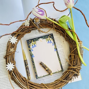 May include: A decorative wreath made of twigs surrounds a notepad and two pens. The notepad has a floral border with blue flowers. Two tulips with purple and white petals and small glass bottles are also in the image.