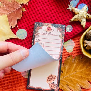 K&ouml;nnte beinhalten: Ein dekorativer Notizblock f&uuml;r Buchrezensionen mit einem skurrilen Design. Der Notizblock hat einen floralen Rand, eine Illustration einer l&auml;chelnden Tasse und die Worte "Book Review". Der Notizblock wird mit einer Seite gezeigt, die angehoben wird und einen leeren Bereich f&uuml;r Notizen freigibt.