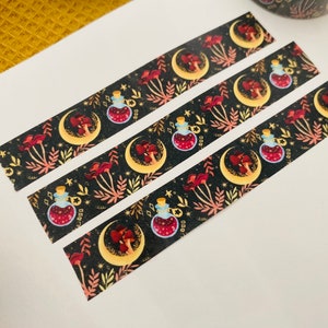 Op de afbeelding: Drie rollen washi tape met een zwarte achtergrond en een herhalend patroon van gouden halve manen, rode paddenstoelen en glazen flessen met een rode vloeistof. De flessen hebben gouden doppen en zijn omringd door gouden sterren en bladeren.