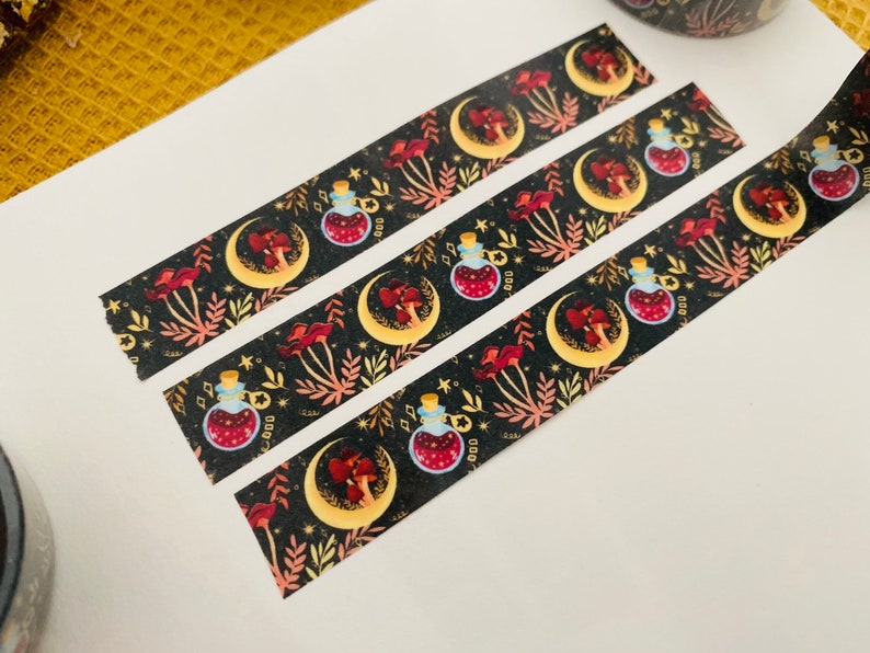 Op de afbeelding: Drie rollen washi tape met een zwarte achtergrond en een herhalend patroon van gouden halve manen, rode paddenstoelen en glazen flessen met een rode vloeistof. De flessen hebben gouden doppen en zijn omringd door gouden sterren en bladeren.