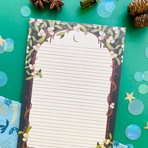 May include: Lined notepad with a decorative border featuring a tree branch design with white berries and green leaves. The notepad has a crescent moon at the top and is surrounded by star anise, a small bottle, and iridescent confetti on a green background.