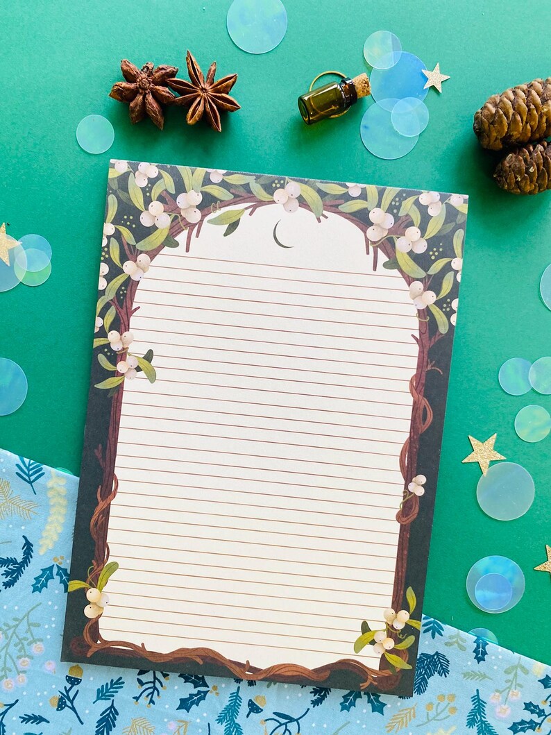 May include: Lined notepad with a decorative border featuring a tree branch design with white berries and green leaves. The notepad has a crescent moon at the top and is surrounded by star anise, a small bottle, and iridescent confetti on a green background.
