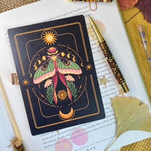 Peut inclure: Un carnet noir avec une bordure dor&eacute;e et une illustration color&eacute;e d'un papillon de nuit avec une lune et des &eacute;toiles. Le texte "The Bamboo-Cutter and the Moon-Child" est imprim&eacute; en bas du carnet.