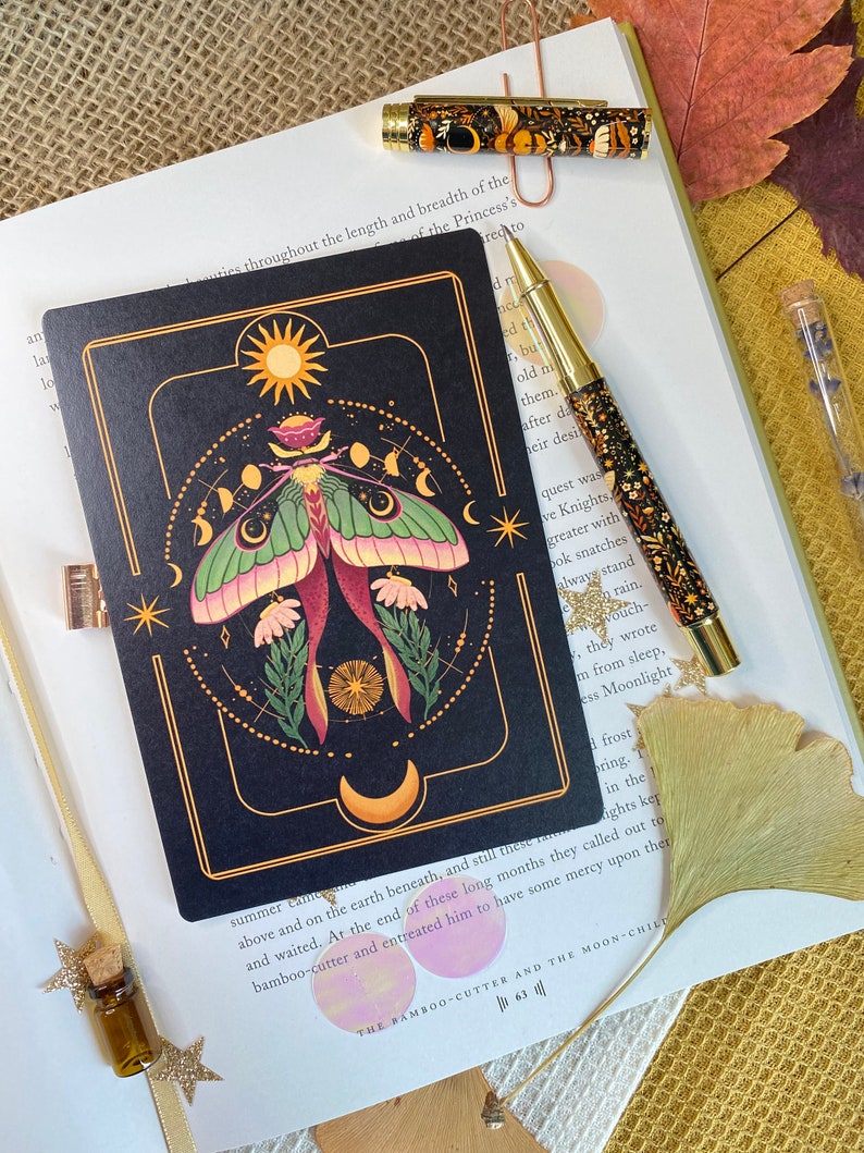 Peut inclure: Un carnet noir avec une bordure dor&eacute;e et une illustration color&eacute;e d'un papillon de nuit avec une lune et des &eacute;toiles. Le texte "The Bamboo-Cutter and the Moon-Child" est imprim&eacute; en bas du carnet.