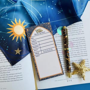 Puede incluir: Un bloc de notas de reseña de libro, un bolígrafo y una decoración en forma de estrella descansan sobre un libro abierto. Una bufanda de seda azul con un diseño de sol y estrella está en el fondo. El bolígrafo tiene un plumín dorado y un barril estampado.
