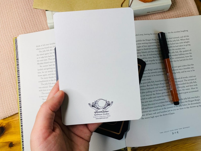 Peut inclure: Une carte blanche avec une illustration en noir et blanc d'une pieuvre stylis&eacute;e avec un cercle autour. Le texte "Splatoon Studios Collection 2023" est imprim&eacute; sous l'illustration.