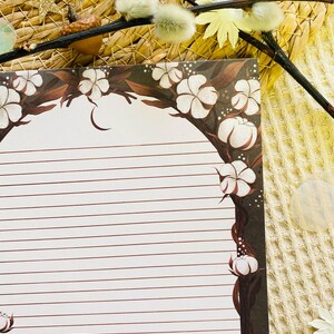 Pu&ograve; includere: Carta da lettere a righe con un bordo floreale di fiori di cotone bianchi e steli marroni. La carta ha uno sfondo bianco con linee orizzontali marroni. Il bordo &egrave; marrone scuro con punti bianchi.