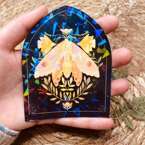 K&ouml;nnte beinhalten: Ein holografischer Aufkleber mit einer Motte mit goldenen und gelben Fl&uuml;geln, umgeben von goldenen Bl&auml;ttern und Blumen auf einem dunkelblauen Hintergrund.