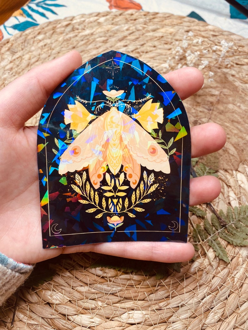 K&ouml;nnte beinhalten: Ein holografischer Aufkleber mit einer Motte mit goldenen und gelben Fl&uuml;geln, umgeben von goldenen Bl&auml;ttern und Blumen auf einem dunkelblauen Hintergrund.