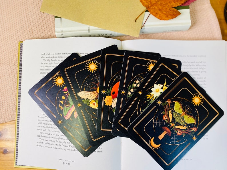 Peut inclure: Un jeu de six cartes de tarot noires avec des accents dor&eacute;s et des illustrations d'insectes, de fleurs et de corps c&eacute;lestes. Les cartes sont dispos&eacute;es en &eacute;ventail sur un livre.