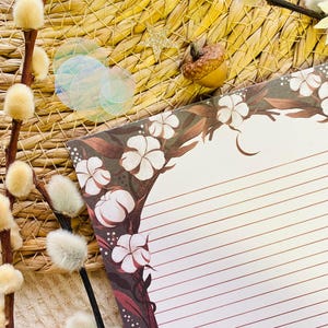 Pu&ograve; includere: Un set di cancelleria decorativo con un blocco note a righe con un design floreale ad albero in marrone e bianco. L'immagine include rametti di salice, cerchi iridescenti, una stella d'oro e una ghianda, il tutto disposto su uno sfondo tessuto.