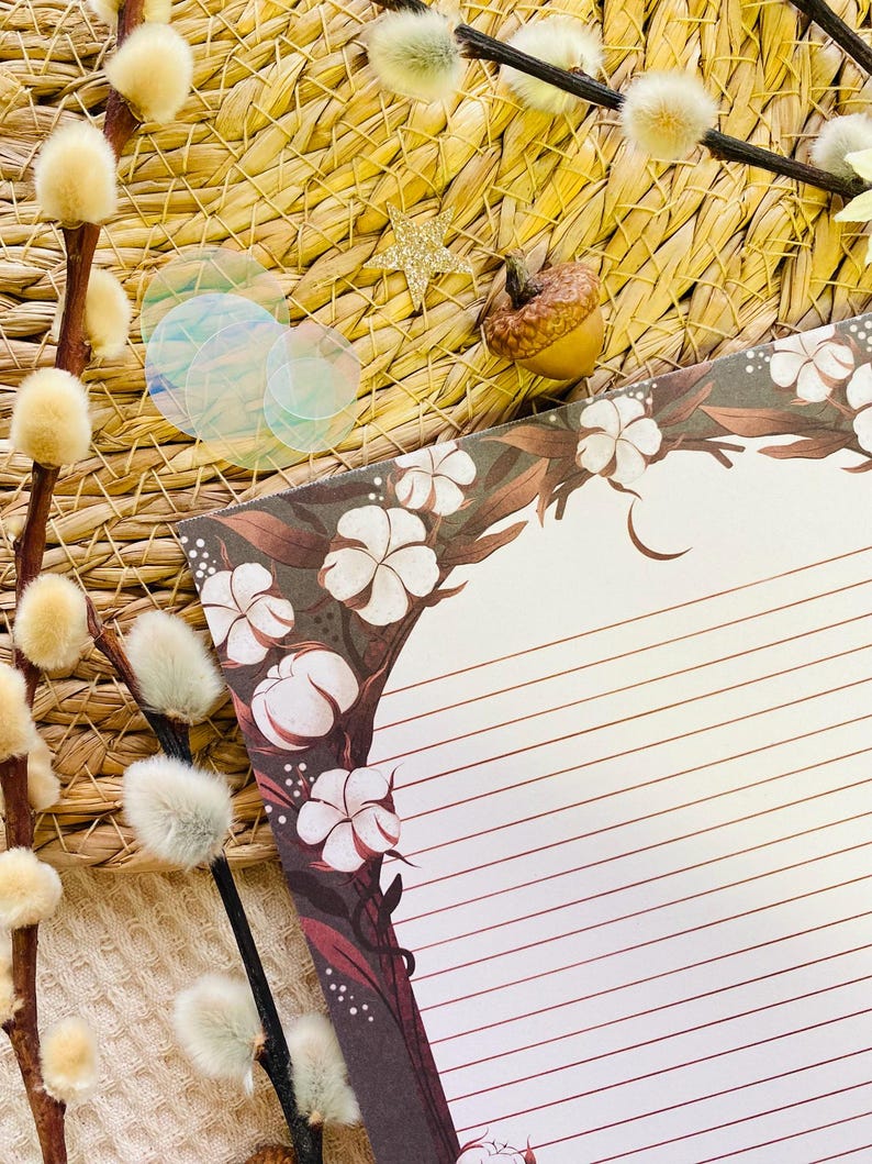 Pu&ograve; includere: Un set di cancelleria decorativo con un blocco note a righe con un design floreale ad albero in marrone e bianco. L'immagine include rametti di salice, cerchi iridescenti, una stella d'oro e una ghianda, il tutto disposto su uno sfondo tessuto.