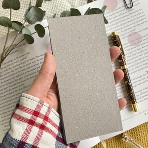 May include: A rectangular, light gray card held in a hand, with a book and pen in the background. The card is made of a textured, matte material. The pen has a gold tip and a dark brown pattern. Eucalyptus sprigs and a book are also visible.