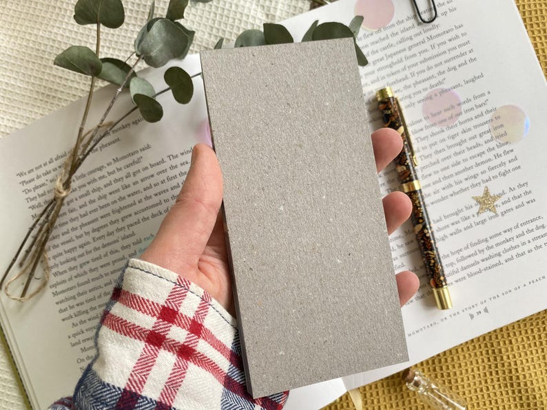 May include: A rectangular, light gray card held in a hand, with a book and pen in the background. The card is made of a textured, matte material. The pen has a gold tip and a dark brown pattern. Eucalyptus sprigs and a book are also visible.
