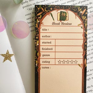 May include: A decorative bookmark with a book review template. The design features a book illustration, pen, and botanical elements. The bookmark includes fields for title, author, and rating.