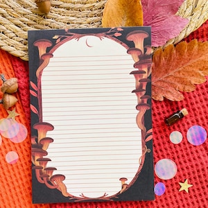 May include: A lined notepad with a black border featuring a mushroom design in shades of brown and orange. The notepad has a white interior with horizontal lines. The background includes autumn leaves, acorns, and decorative elements.