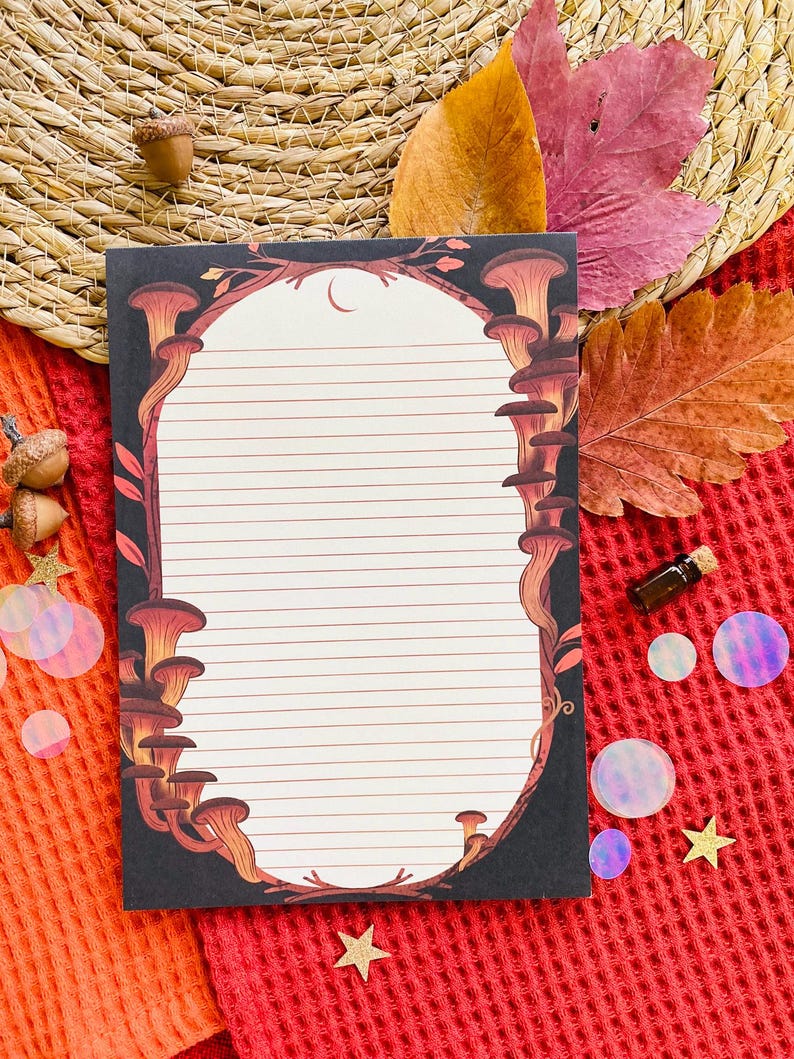 May include: A lined notepad with a black border featuring a mushroom design in shades of brown and orange. The notepad has a white interior with horizontal lines. The background includes autumn leaves, acorns, and decorative elements.