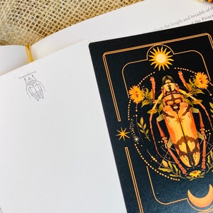 May include: A black card with a gold border features a detailed illustration of a beetle with a sun and moon in the background. The card also includes the text "Tmesisternus" and "Karen Dubas Art Studios 2024".