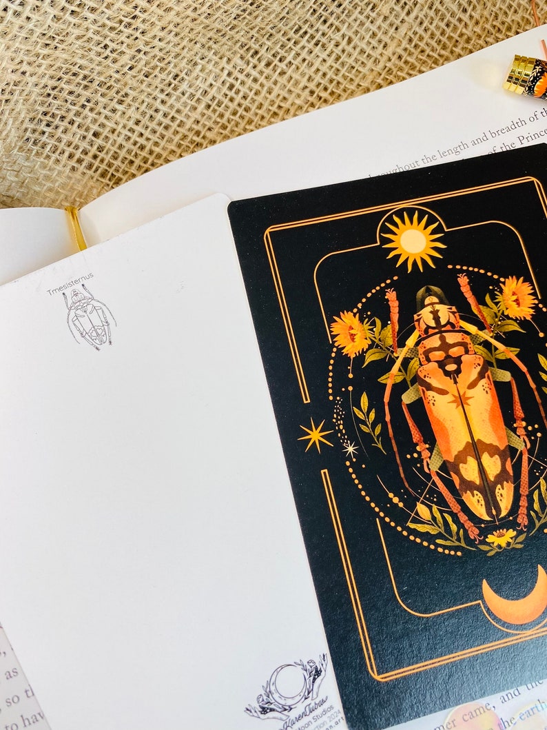 May include: A black card with a gold border features a detailed illustration of a beetle with a sun and moon in the background. The card also includes the text "Tmesisternus" and "Karen Dubas Art Studios 2024".