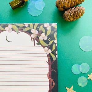May include: An overhead shot features a green background with a lined notepad, a small glass bottle with a cork, two pine cones, and decorative elements. The notepad has a botanical border with white berries and a crescent moon.