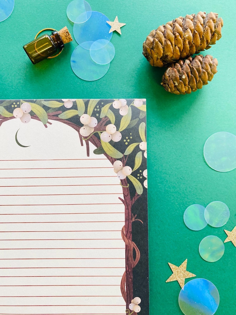 May include: An overhead shot features a green background with a lined notepad, a small glass bottle with a cork, two pine cones, and decorative elements. The notepad has a botanical border with white berries and a crescent moon.